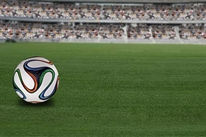Top i bardhë me dizajne shumëngjyrëshe mbi një fushë gjelbër futbolli me tribuna të mbushura me spektatorë