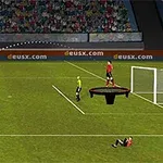 Një lojtar futbolli godet një top drejt portës, ndërsa një lojtar në një platformë qëndron pranë portës dhe një tifoz është në tribunë